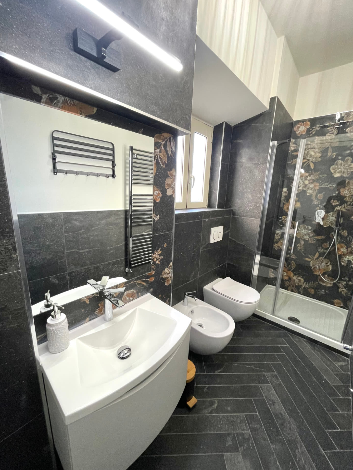 Modern bathroom at Dolce Apartments with dark tiling, white sink and floral-tiled walk-in shower